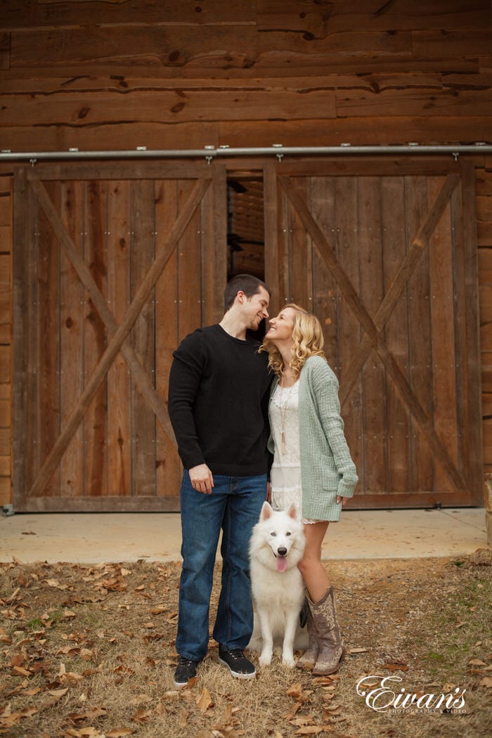 In this photograph, the couple smiles in front of rustic barn doors that are made of natural wood. They are not alone though, they are with their beloved companion and best friend.