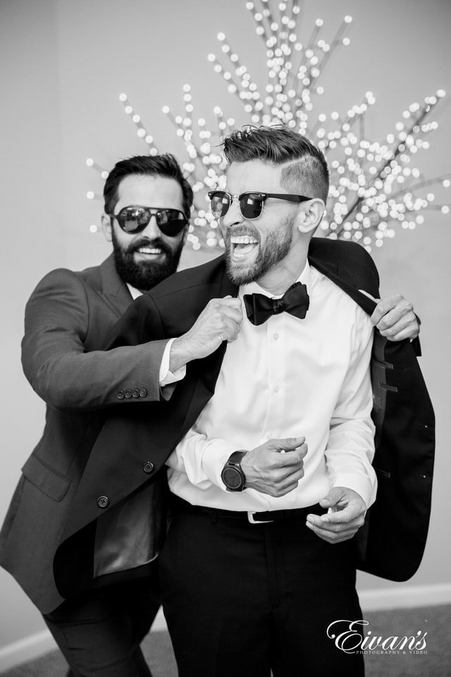 Black and white photo of a groom laughing with his groomsman as he shrugs into his tuxedo jacket.