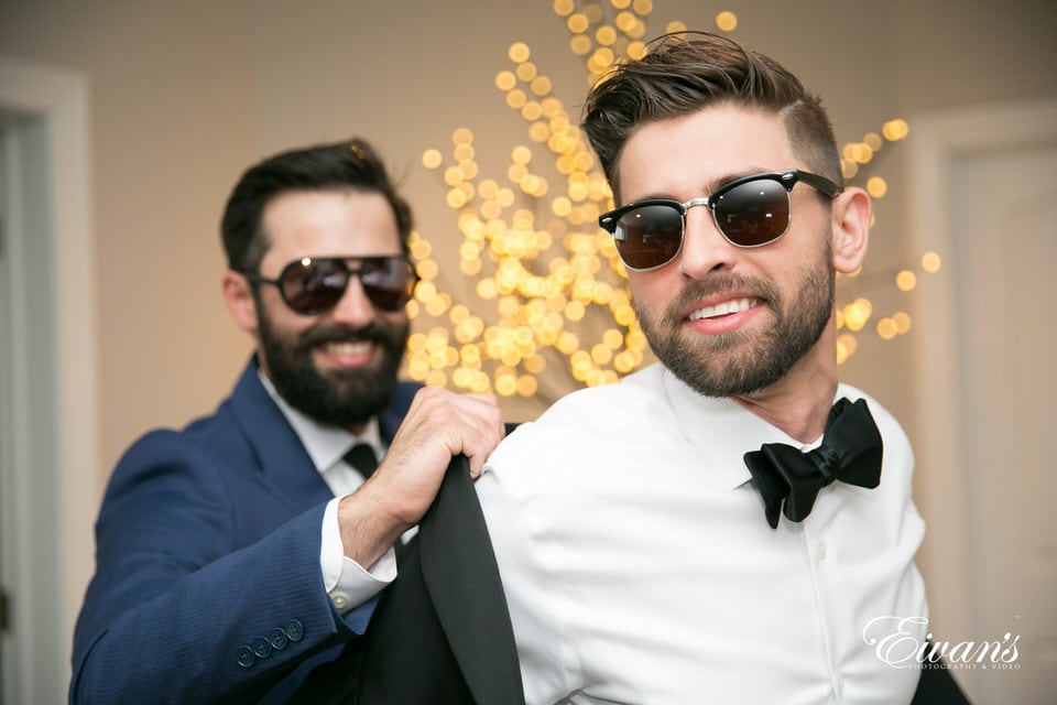 The groom gets help putting his tuxedo jacket on by his groomsman. They both sport dark sunglasses and kind smiles.