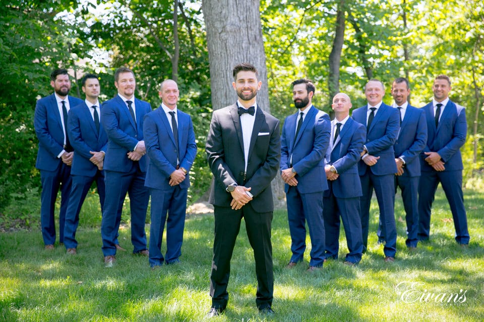 A groom stands, smiling with his arms crossed at the wrist in a forest-like area. His groomsmen flank him on either side in a diagonally increasing line with arms crossed at the wrist.