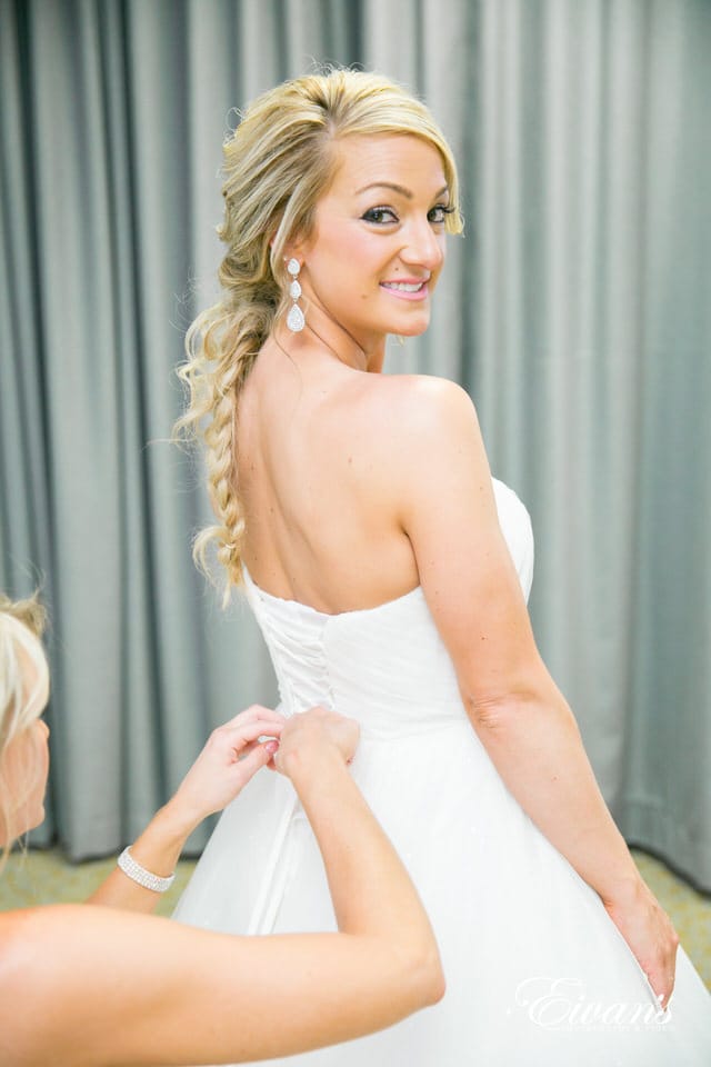 A happy bride looks over her shoulder at the photographer. Her smile is broad and highlighted with pink lips and 3 tier drop earrings. Her blonde hair is tangled in a loose fishtail braid with escaping tendrils. One of her bridesmaids is carefully lacing up the back of her wedding dress.