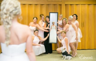 The photographer captures a reflective photo of a bride smiling and looking into a rectangular mirror held by her excited bridal party. Her small, tuxedo-clad dog sits sweetly alongside the bridesmaids.