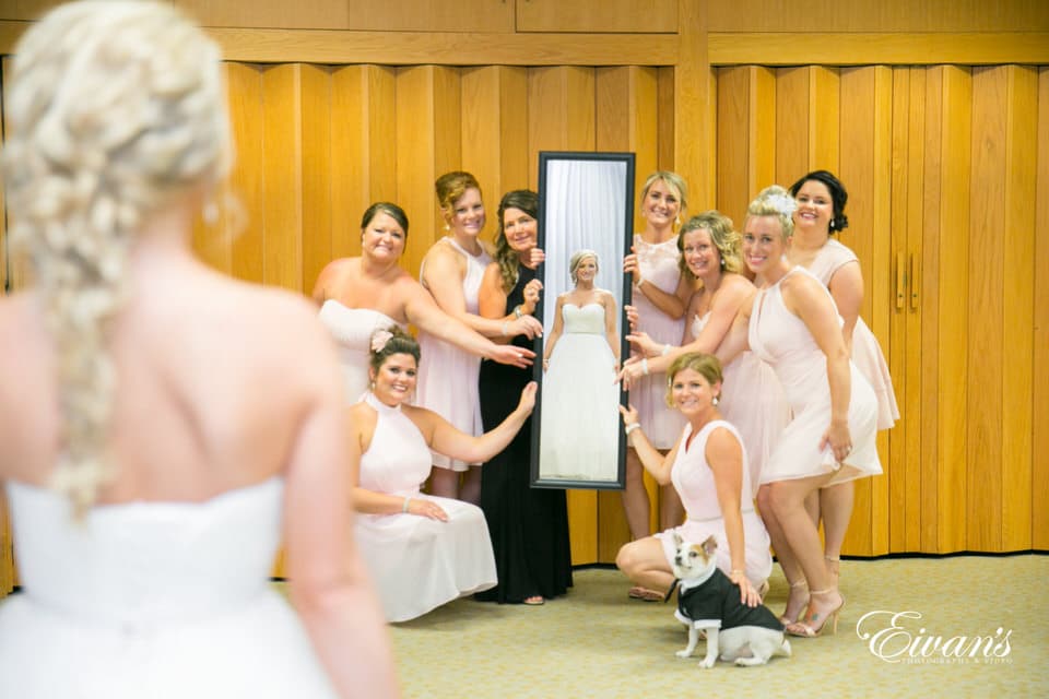 The photographer captures a reflective photo of a bride smiling and looking into a rectangular mirror held by her excited bridal party. Her small, tuxedo-clad dog sits sweetly alongside the bridesmaids.