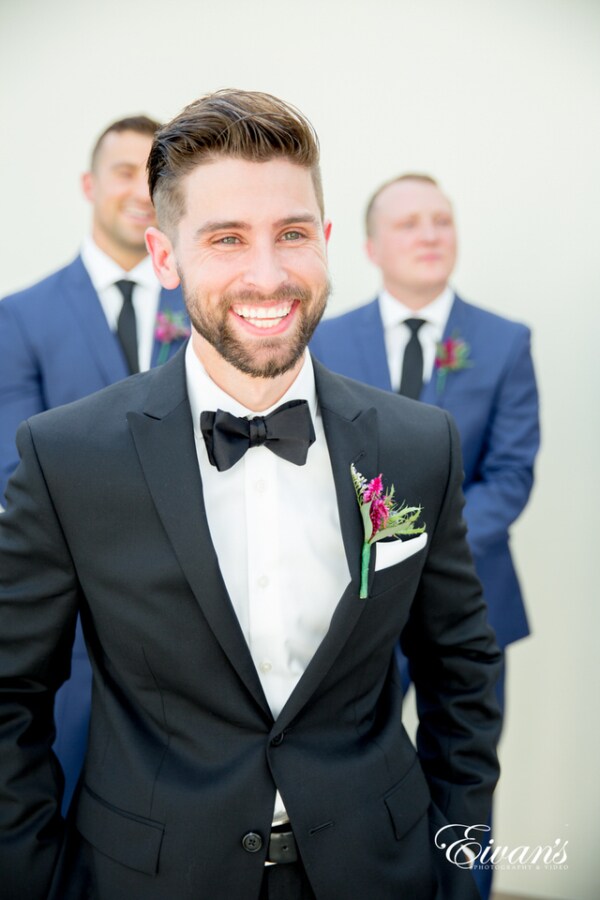 Amelia & Farron Gaerte A groom smiles widely for the photographer as his groomsmen stand smiling behind him. The groom wears a black satin bowtie and a wildflower boutonniere.
