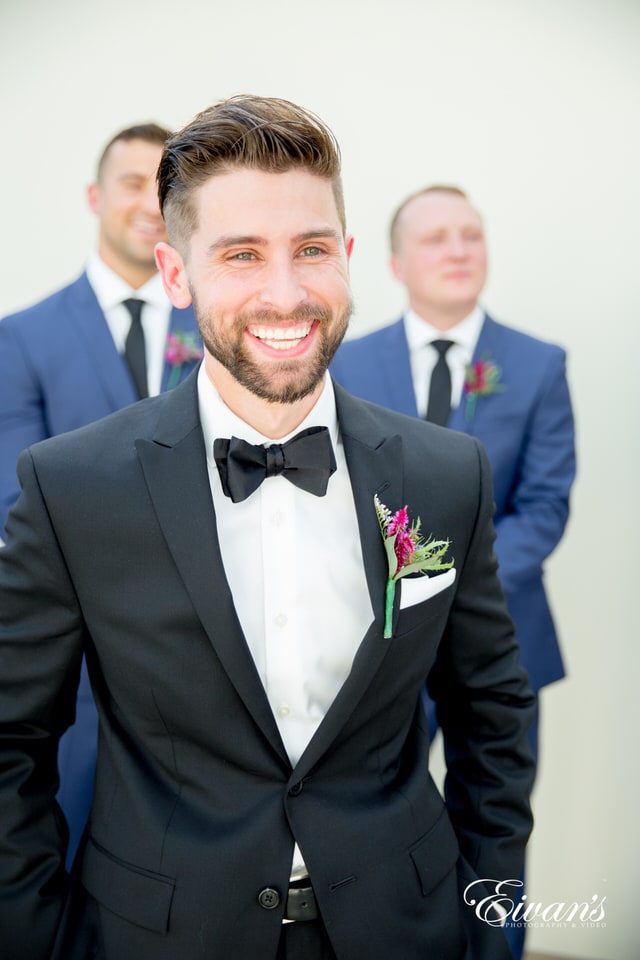 A groom smiles widely for the photographer as his groomsmen stand smiling behind him. The groom wears a black satin bowtie and a wildflower boutonniere.