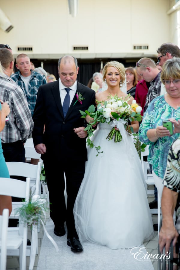 Amelia & Farron Gaerte A bride smiles widely as her father walks her down the aisle while guests prepare their phone cameras to take pictures.