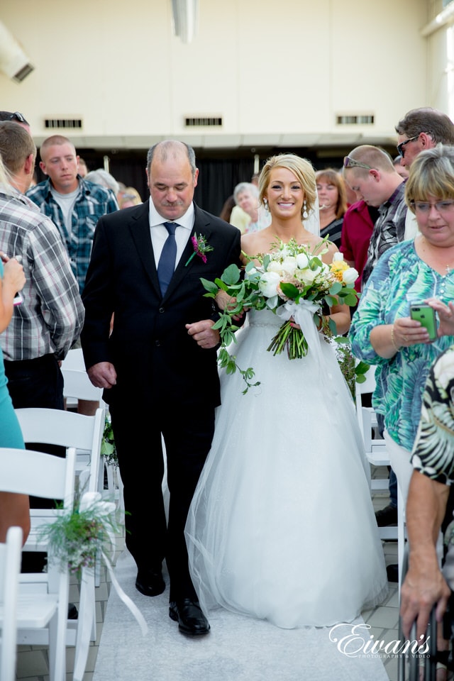 A bride smiles widely as her father walks her down the aisle while guests prepare their phone cameras to take pictures.