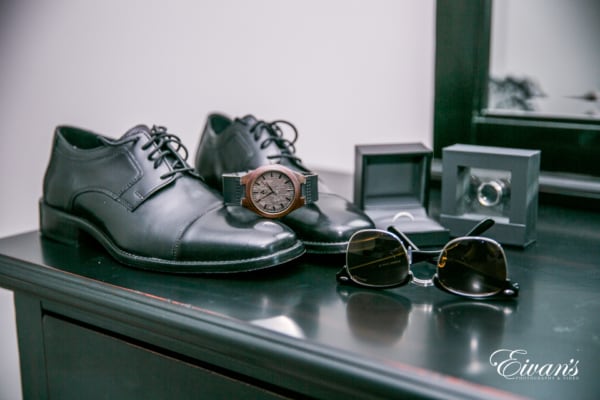 Amelia & Farron Gaerte The photographer captures a detail shot of the groom's shoes, watch, sunglasses and rings as they sit on the corner of a dresser.
