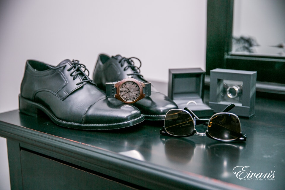 The photographer captures a detail shot of the groom's shoes, watch, sunglasses and rings as they sit on the corner of a dresser.