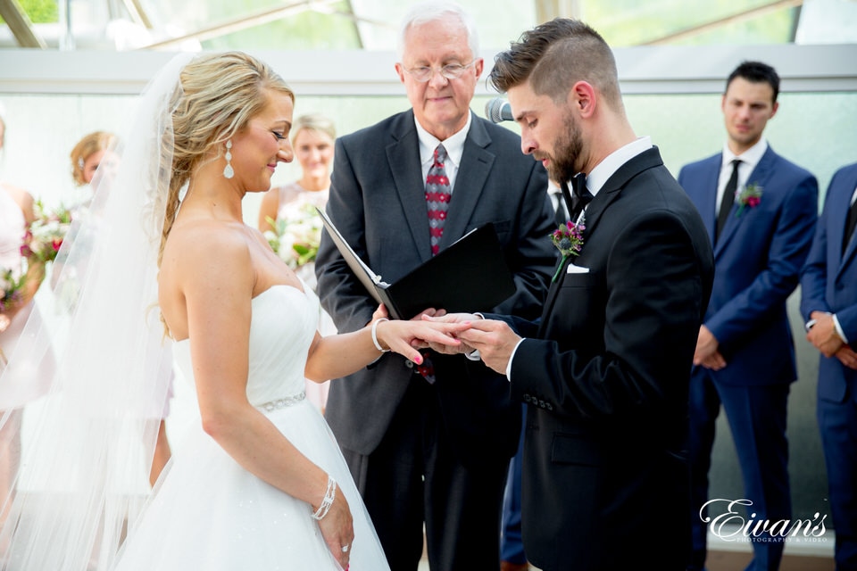 A groom concentrates on putting the wedding band on his bride's finger during their wedding ceremony as the officiant, and bridal party eagerly watch. The bride smiles as she watches her groom slide the ring on her finger.