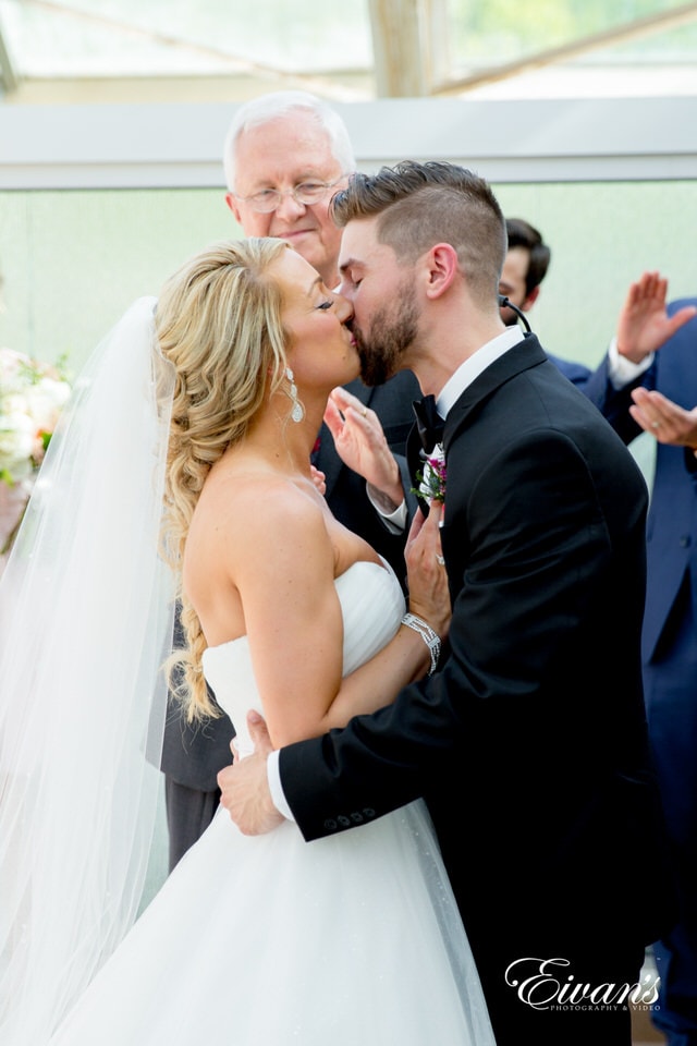 A bride and groom have their first kiss as man and wife at the end of their ceremony. The officiant smiles subtly, and surrounding guests stand and clap.