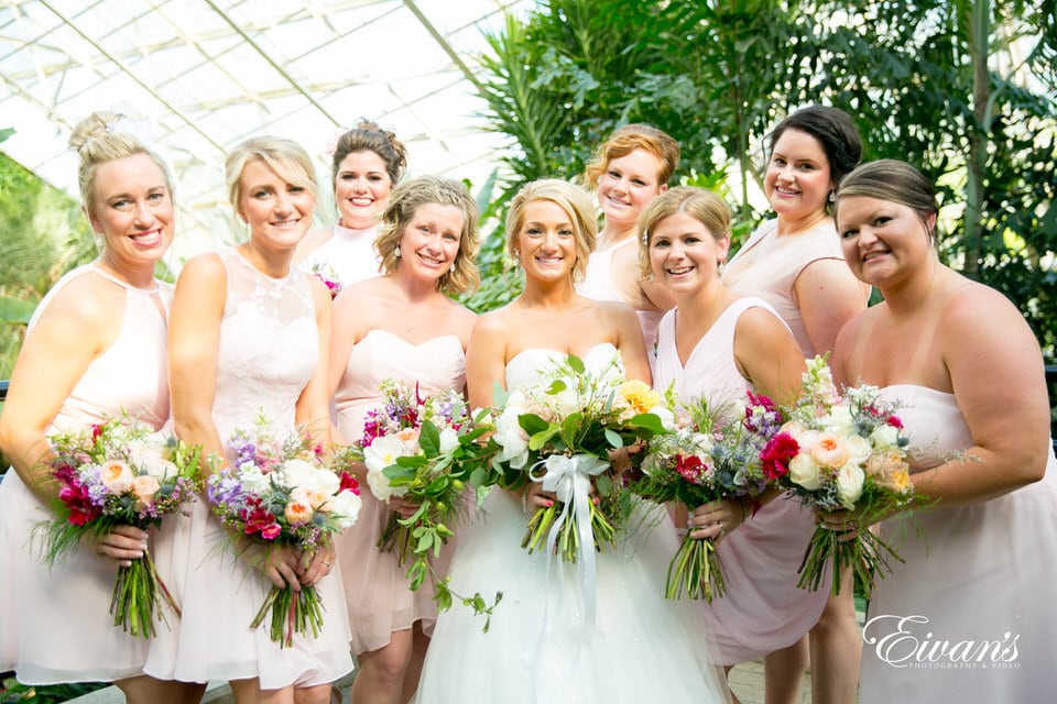 A bride smiles widely, flanked by her bridesmaids with their bouquets in hand. Greenery and tall trees surround the group inside of a high-ceiling glass shelter.