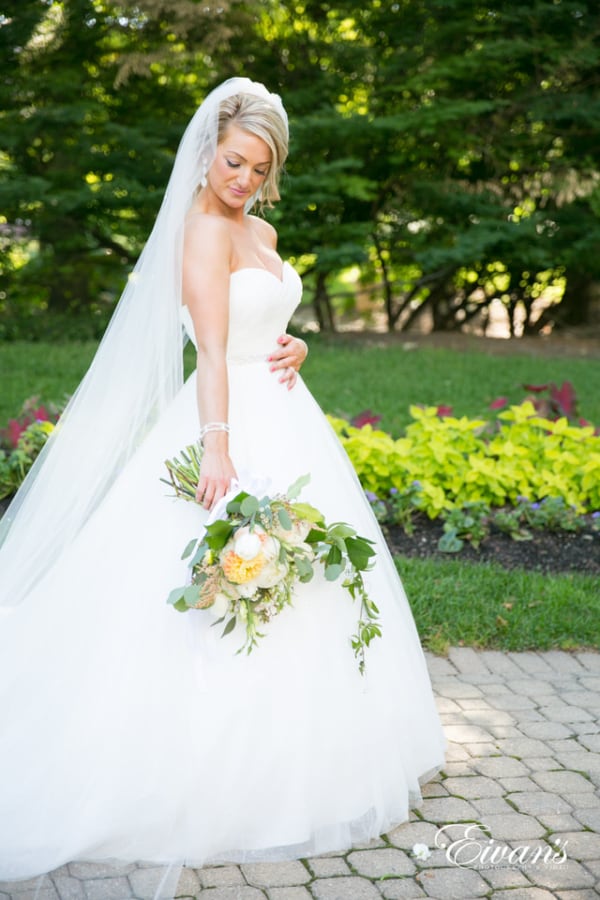 Amelia & Farron Gaerte A bride admires her bouquet while walking through the garden wearing a floor-length veil.