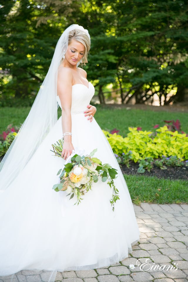 A bride admires her bouquet while walking through the garden wearing a floor-length veil.