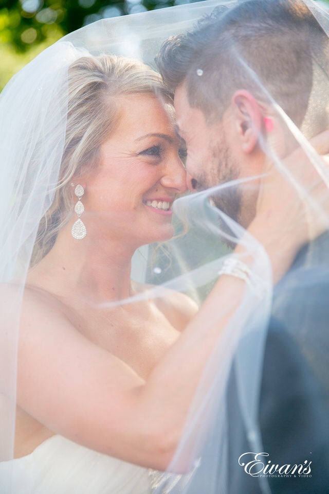 A bride and groom smile and look at one another under the cover of the bride's veil.