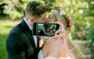 The photographer takes a portrait of a bride snapping a selfie of her and her new husband sharing a kiss.