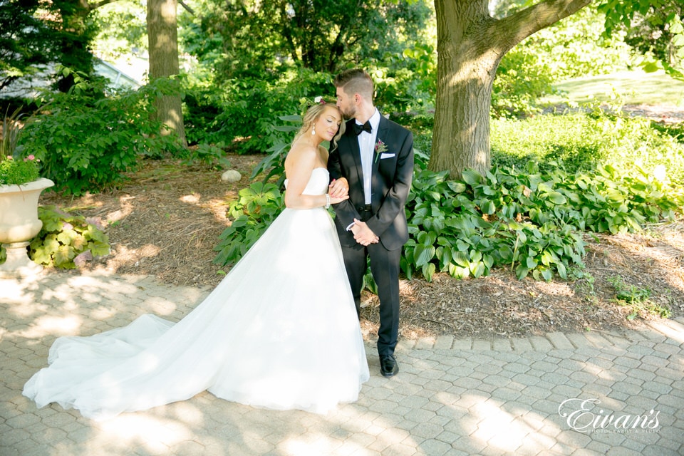 A bride and groom stand in a garden area as they pose for portraits. The groom kisses the forehead of his bride as she rests her head on his shoulder.