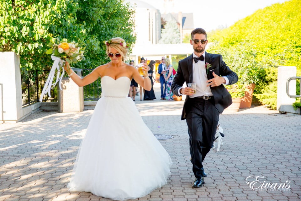 The bride and groom playfully walk toward the photographer as their little dog trails behind them wearing sunglasses with drinks in hand.