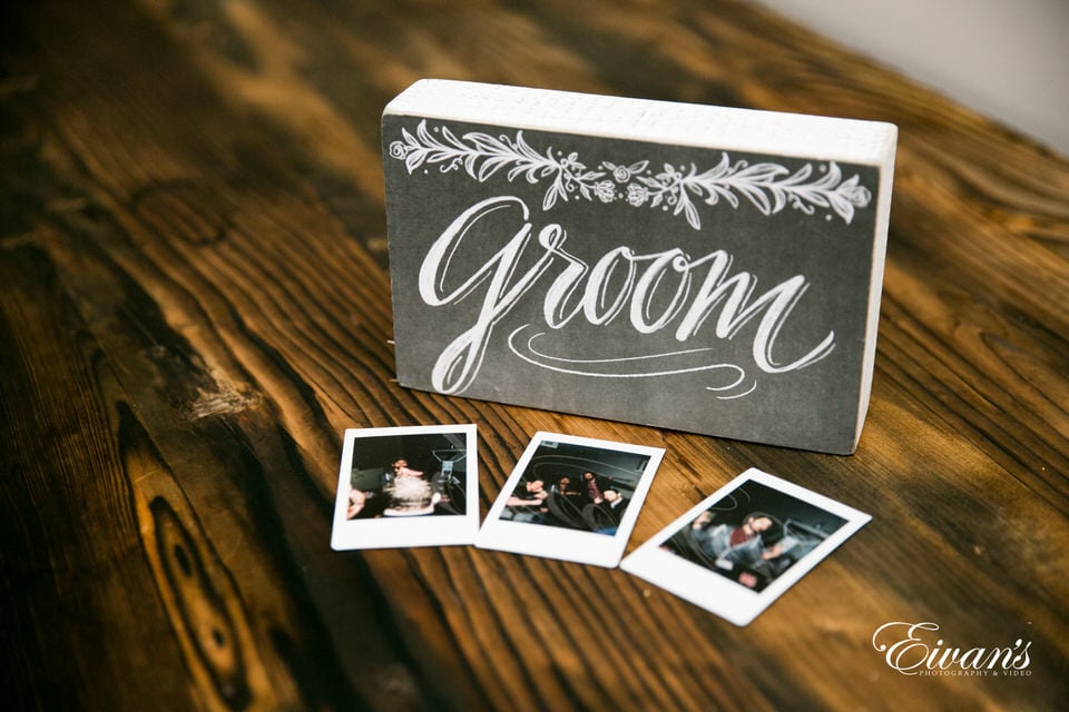 A small, decorative block with the word "groom" printed on the front rests atop a vintage wood table. Three small polaroid photos lay in front of it.