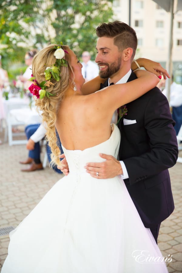 Amelia & Farron Gaerte The groom smiles at his new wife as they dance together in a courtyard with their guests lingering in the background. The bride wears a simple hot pink, green and ivory floral crown.