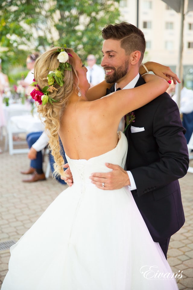 The groom smiles at his new wife as they dance together in a courtyard with their guests lingering in the background. The bride wears a simple hot pink, green and ivory floral crown.