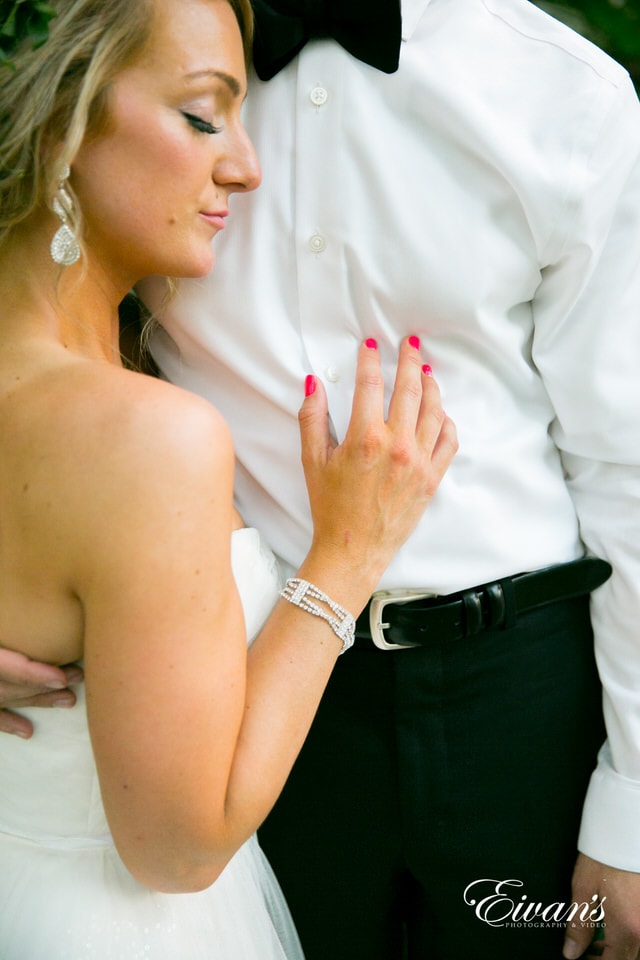 The groom embraces his bride as she stands, resting her head against his chest. The bride sports hot pink nails and silver, jeweled jewelry.