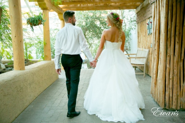 Amelia & Farron Gaerte The photographer captures the bride and groom walking away, hand-in-hand and looking at each other on a back porch shaded from the sun. The bride shows off her loose fishtail hairstyle with a simple flower crown.