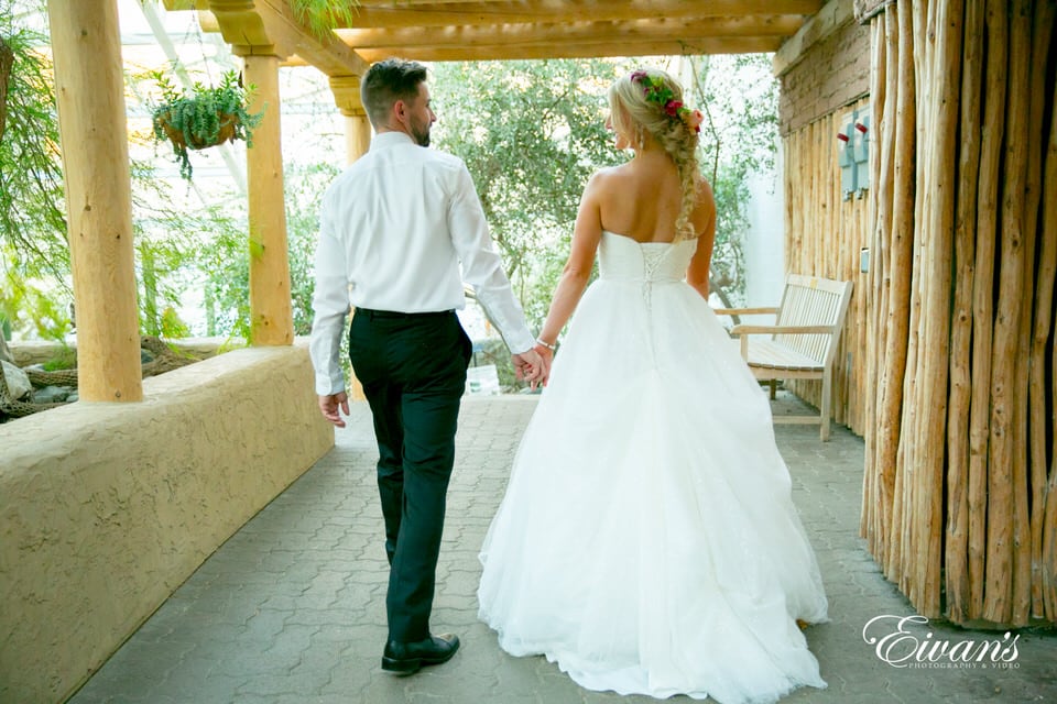 The photographer captures the bride and groom walking away, hand-in-hand and looking at each other on a back porch shaded from the sun. The bride shows off her loose fishtail hairstyle with a simple flower crown.