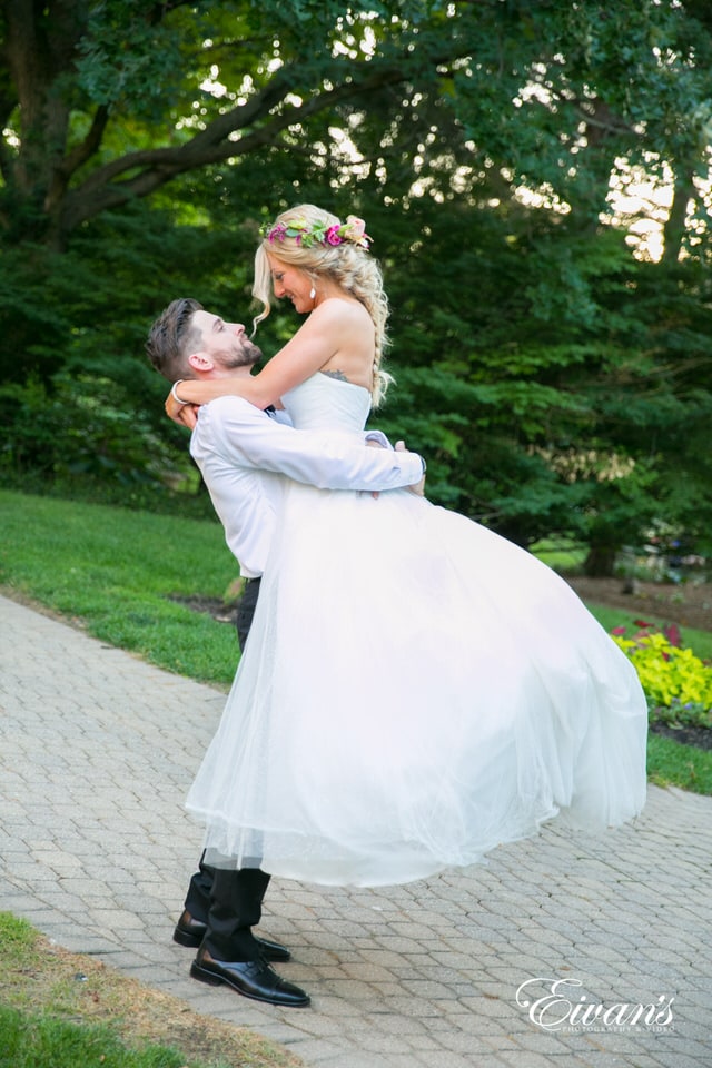 Spellbound groom romantically lifts his new bride into the air. The bride sports a bohemian look with a hot pink flower crown and a fishtail braid hairstyle.