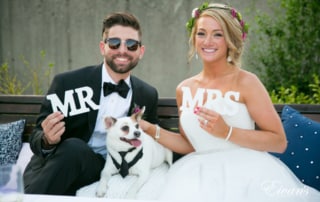 Newlyweds proudly hold their MR and MRS signs as they pose for a photo with their little dog in a tuxedo.