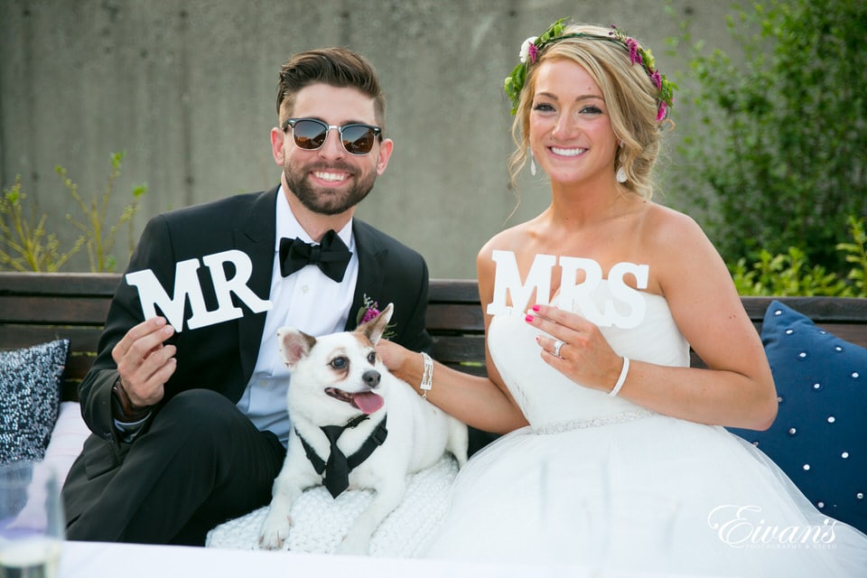 Newlyweds proudly hold their MR and MRS signs as they pose for a photo with their little dog in a tuxedo.