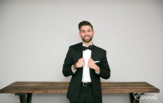 The groom grips the lapels of his tuxedo jacket and smiles broadly in front of a vintage wood table.