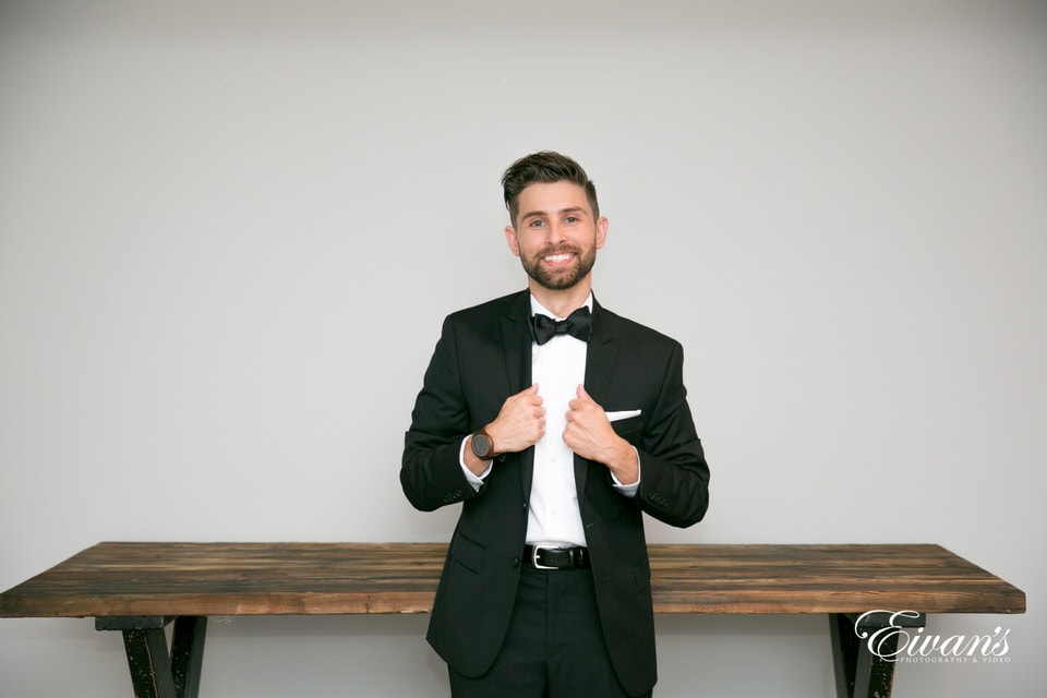 The groom grips the lapels of his tuxedo jacket and smiles broadly in front of a vintage wood table.