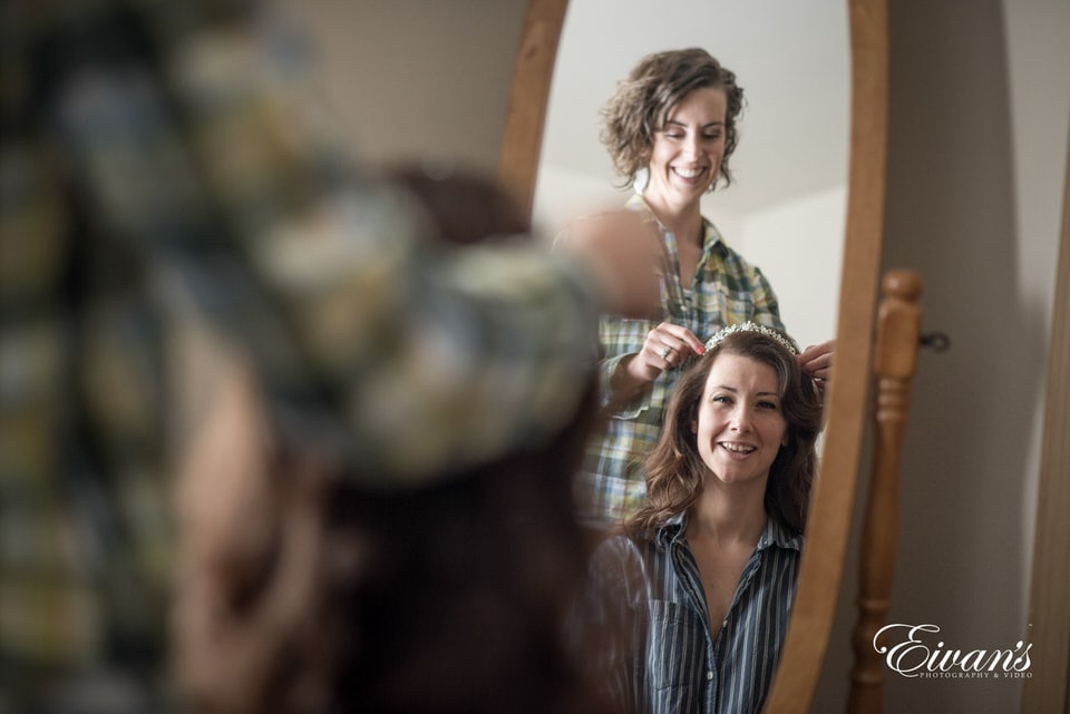 The mother of the bride joyfully helps her daughter get ready by placing her tiara as they smile and laugh in the mirror.