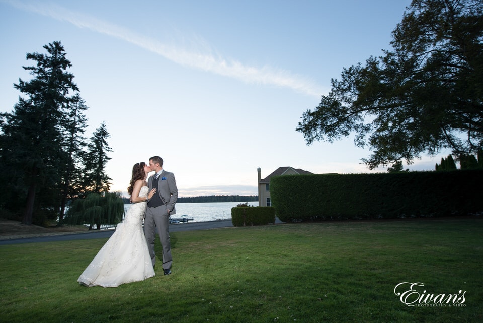 Newlyweds share a kiss on an open, grassy field overlooking a body of water at sunset.