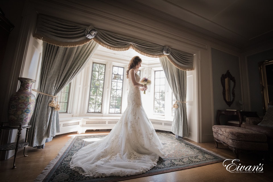 Shawna & Steven The photographer takes a stunning image of the bride looking thoughtfully at her bouquet in the middle of a vintage style room with deep bay windows. Her long train spills out on the floor beneath her.