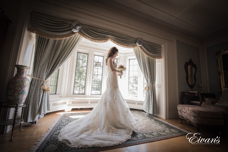 The photographer takes a stunning image of the bride looking thoughtfully at her bouquet in the middle of a vintage style room with deep bay windows. Her long train spills out on the floor beneath her.