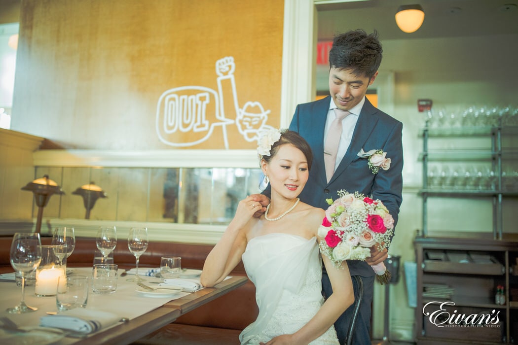 The groom hands his bride a white and pink rose bouquet as she sits of so elegantly at a lovely table.