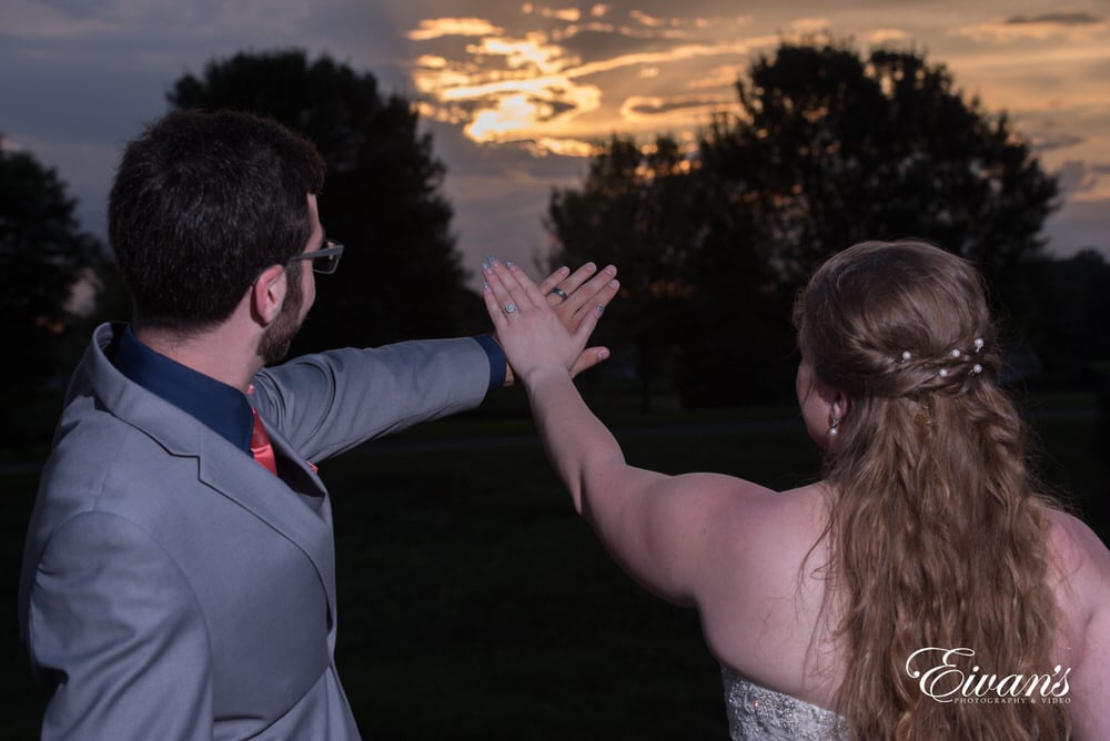 The couple hold their hands up showing off their rings into the sunset.