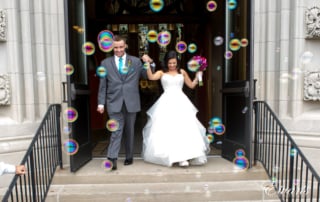 The couple smiles and gleams while they walk out of the chapel while bubbles are being blown at them.