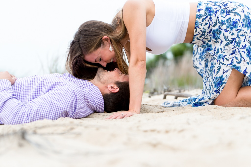 A newly engaged woman playfully hangs her head over her soon-to-be husband as he lies in the sand of the beach.