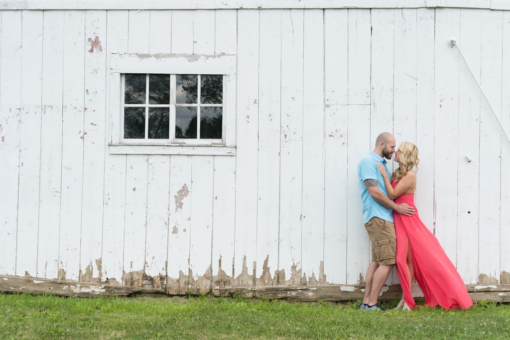 A newly engaged couple looking adoringly at one another in front of an old, rustic barn.