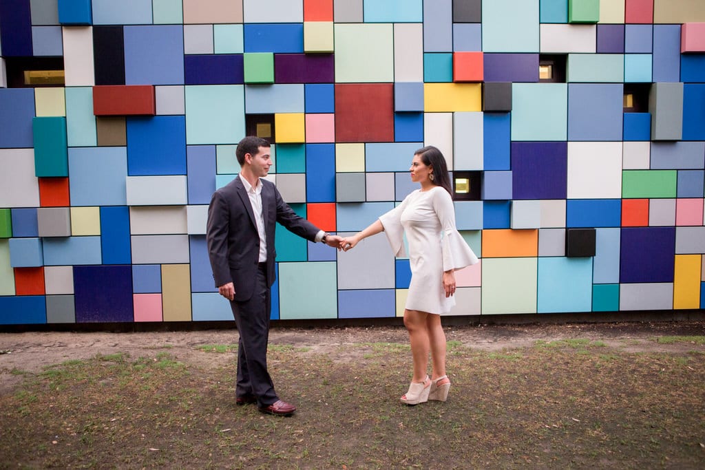 A newly engaged couple stand face to face holding hands in front of a multi-colored, geometric wall.