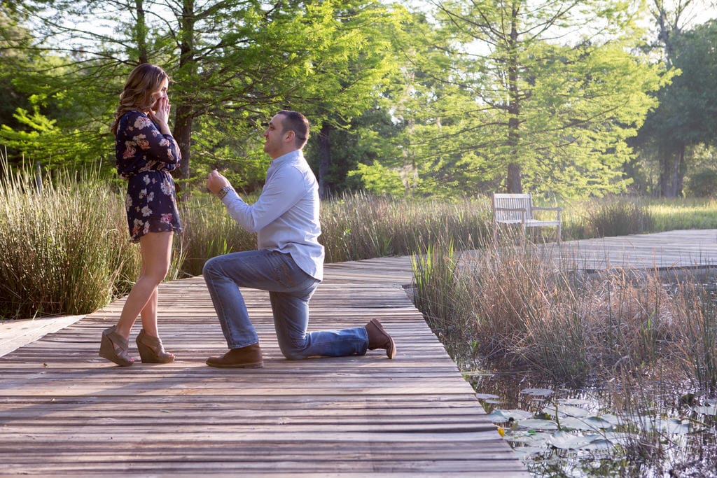 A man proposes to the girl of his dreams as she excitedly accepts. He is kneeling on a beautiful, wooden bridge holding the ring before her on a gorgeous sunny day in the forest.