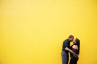Wedding / Engagement Photography A newly engaged couple, clad in all black, share a kiss in front of a yellow background.