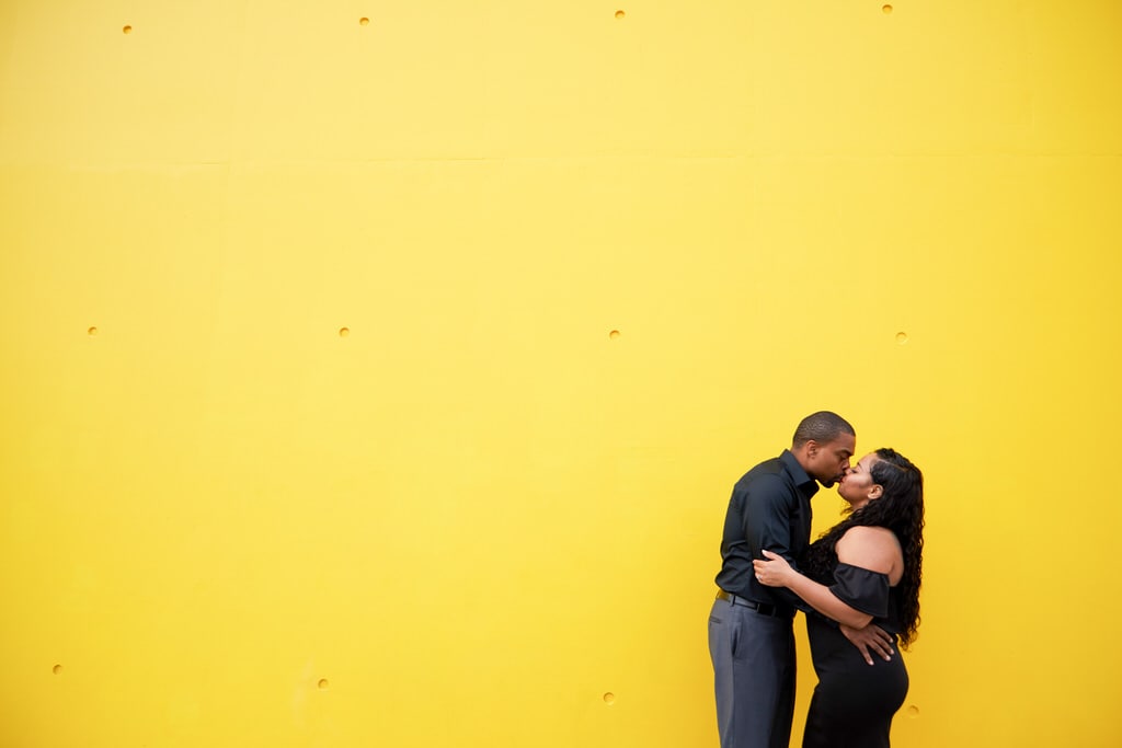 A newly engaged couple, clad in all black, share a kiss in front of a yellow background.