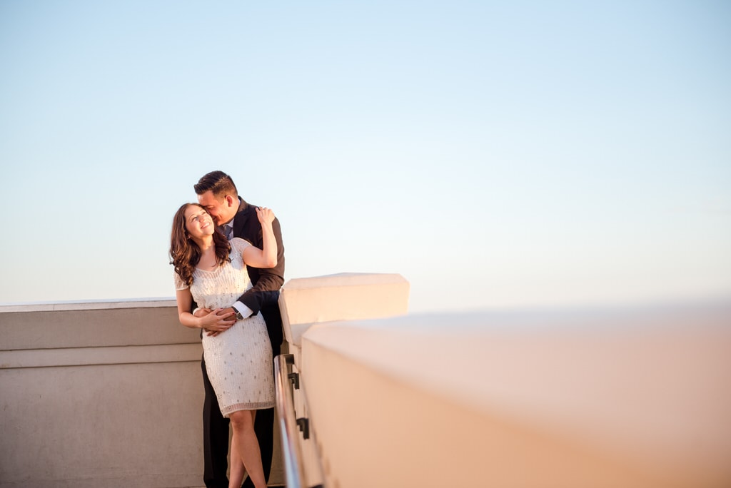 A man embraces his soon-to-be wife on a roof in the late afternoon. He kisses her head, and she smiles widely while grabbing his shoulder.