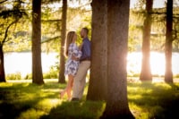 Abbey & Shawn 4/14/18 A newly engaged couple leans against a tree near a body of water. They stand in the grass and smile at each other.