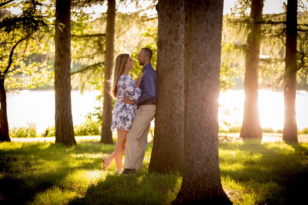 A newly engaged couple leans against a tree near a body of water. They stand in the grass and smile at each other.