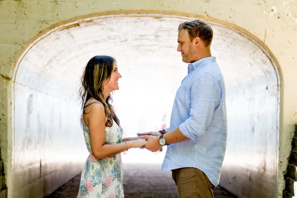 A newly engaged couple hold hands and smile at one another in front of a wide tunnel.